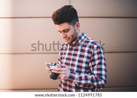 Skilled male photographer in casual stylish shirt viewing pictures on camera satisfied with results, hipster guy  amateour enjoying leisure time on hobby making settings on equipment for taking photo