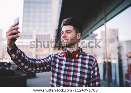Cheerful hipster guy in casual wear making selfie on front camera of modern smartphone spending time outdoors, happy male blogger taking picture for share in networks and update profile via cellular