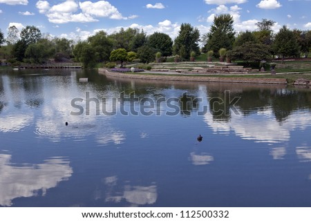 Calm waters in a large pond at the Overland Park (Kansas) Arboretum reflects fair weather cumulus clouds against a blue sky. Royalty-Free Stock Photo #112500332