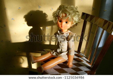 Old vintage blond girl with dark-eyed doll sitting on a chair against a background of wallpaper