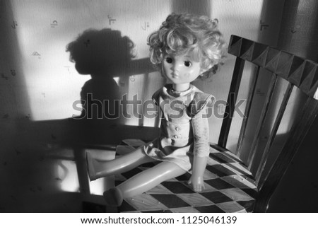 Old vintage blonde girl with a doll with dark eyes, sits on a chair on a black and white background