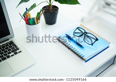 Close up of notebook lying on table with pair of glasses on top. Cup with pens, plant and computer are nearby