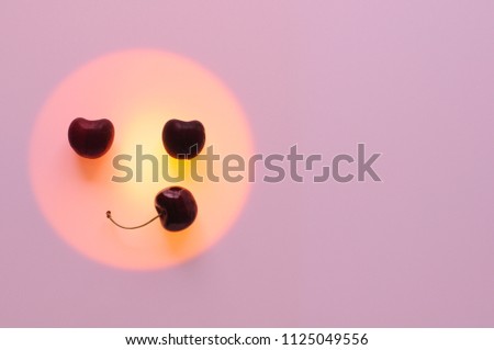 Red cherry fruits put on the orange color circle set as smiling emoji emotion face with pink background that have space for text.