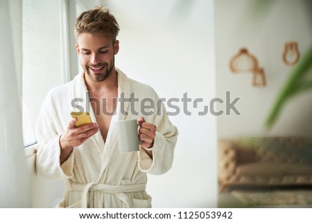 Waist up portrait of satisfied male drinking coffee in morning. He is standing on room holding cup and mobile in hands. He is looking at screen with joy. Copy space in right side