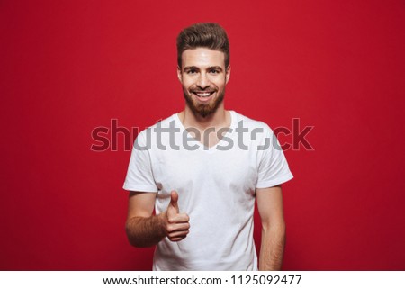 Image of handsome cheerful young man in white t-shirt looking camera showing thumbs up isolated over red background.