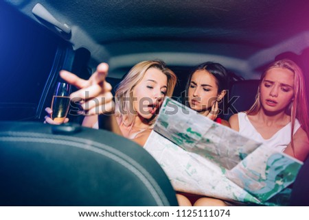 Three girls are sitting in car and looking on map. Blonde girl is pointing forward. They are talking with each other. Another blonde girl is upset. Brunette looks very serious.