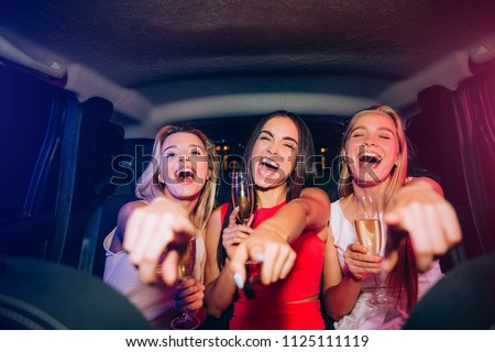 Happy and excited girls are sitting together in car. They are looking and pointing on camera. Young women are laughing and smiling. Some of them has a glass of champagne in hands.