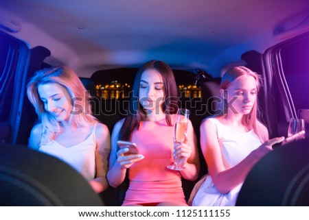Another picture of three beautiful girls are sitting together in car and looking at their phones. Also they are holding glasses of champagne in hands. Girls are concentrated and serious.