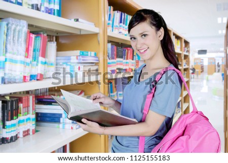 Picture of pretty college student smiling at the camera while reading a textbook and standing in the library