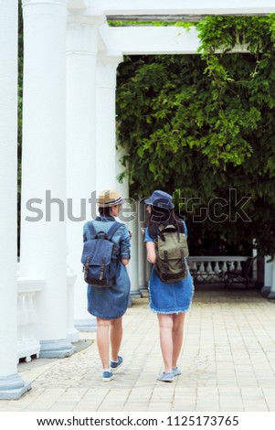 Two young beautiful girls are walking along a green park with white columns. Day, the sun. Travelers, tourists, laugh and take pictures on a smartphone and camera.