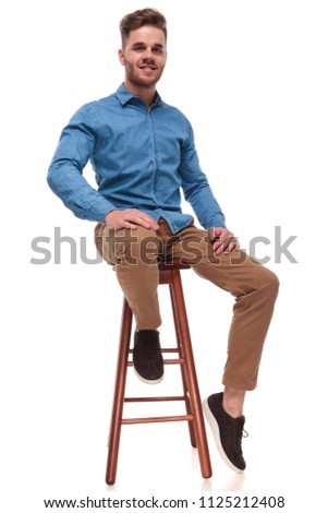 attractive casual man wearing a blue shirt sits on wooden stool on white background, full body picture