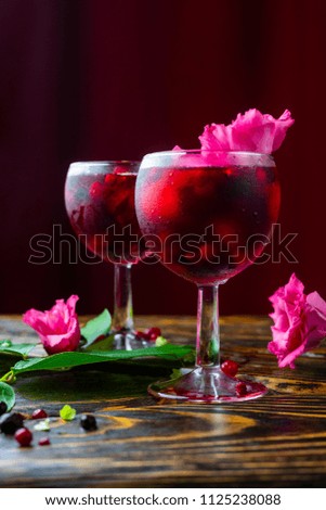 Two glasses full of red drink with berries, ice and a pink flowers, beside which lie berries and a branch of green tropical leaves on a vintage wooden table. Copy space. Commercial design.