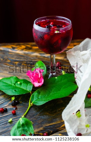 A glass full of red drink with berries, ice and a pink flower on crumpled wrapping paper beside which lie berries and a branch of green tropical leaves on a vintage wooden table. Copy space.