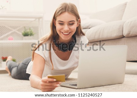 Happy young blonde woman shopping online with credit card and laptop, lying on floor at home, copy space