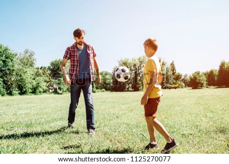 A picture of father nd son playing football game. THere is a ball flying from dad to son. It is in air. It is sunny outside on field.