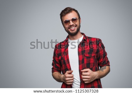 Stylish young man with tattoo and beard wearing casual checkered shirt and sunglasses smiling at camera