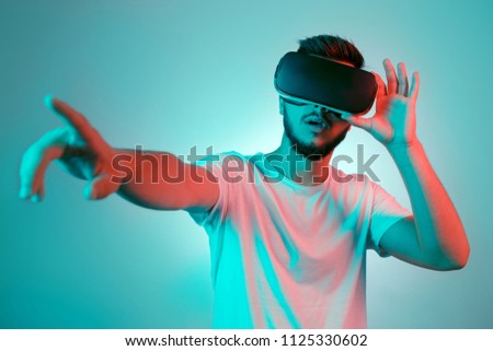Handsome bearded man trying VR headset and poniting. Young man exploring another world with virtual reality goggles on the colorful background.
