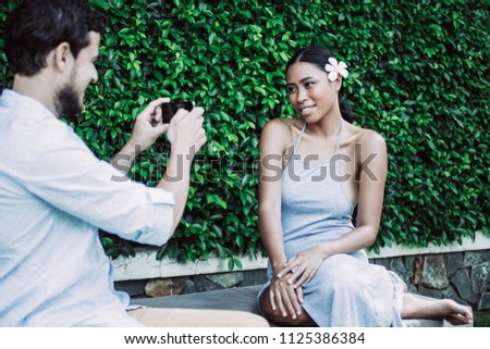 Closeup of Caucasian man taking photo of smiling pretty young Asian woman sitting on chaise longue with green leaves wall in background
