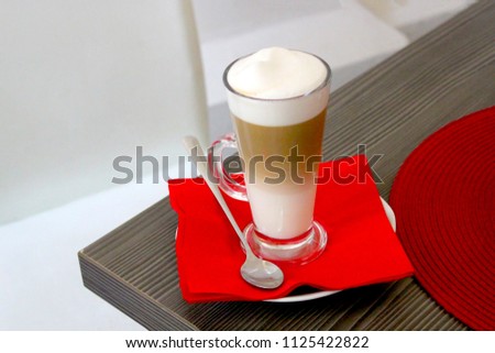 Glass mug with latte on is on the wooden table. There is a red napkin and spoon. The pot is located in the café.