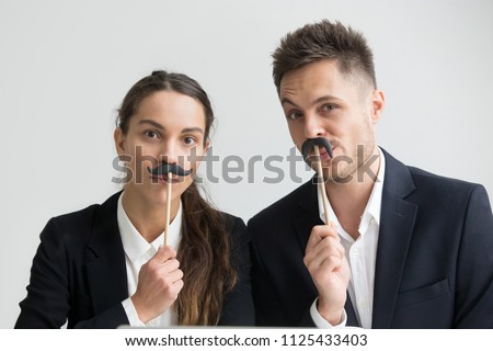 Funny male and female colleagues in suits holding fake mustache, businessman and businesswoman making silly faces, millennial coworkers grimacing looking at camera, creative team head shot portrait