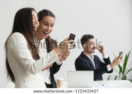 Diverse female colleagues laughing having fun with smartphone in office while male coworker taking silly selfie, excited multi-ethnic business people watching funny online video or using mobile apps