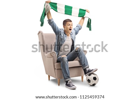 Overjoyed teenage soccer fan with a scarf and a football sitting in an armchair isolated on white background Royalty-Free Stock Photo #1125459374