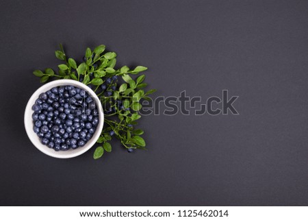 Top view of wild organic blueberries with green leaves in a white bowl on black background with copy space. Flat lay.