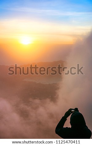 Young man taking pictures the foggy dawn on top of the volcano Batur, Kintamani, Bali island, Indonesia. Sunrise on the volcano of Batur, amazing natural landscape. Travel concept.