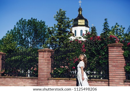 Outdoor portrait of young beautiful curly girl near forged lattice with roses. Photo of girl with beautiful hair and makeup on background of Orthodox Church.