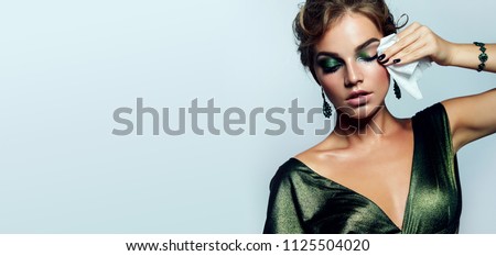 beautiful young girl with a bright make-up and in a shiny green dress striatet makeup from her face with a wet napkin. Hairstyle - curls are gathered in a bun. Royalty-Free Stock Photo #1125504020