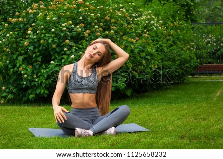 A dark-haired woman coach in a sporty short top and gym leggings does stretching and pulling his head  on the rug for yoga, hands are raised upwards,  on a summer day in a park on a green lawn