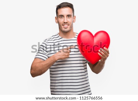 Handsome young man in love holding red heart very happy pointing with hand and finger