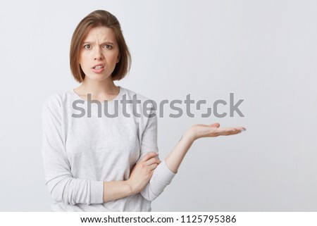 Closeup of embarrassed displeased young woman in longsleeve looks confused and holding copyspace on palm isolated over white background