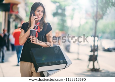 Portrait of woman holding paper bags and coffee on the street after shoping while using smart phone. Royalty-Free Stock Photo #1125813590