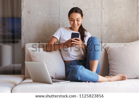 Happy young asian woman using mobile phone while sitting a couch at home with laptop computer Royalty-Free Stock Photo #1125823856