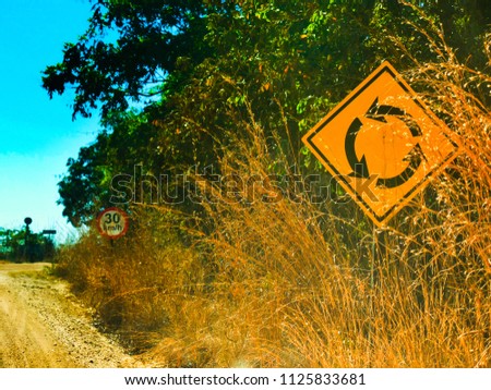 Two traffic signs on a dirt road, with dry vegetation and green vegetation.