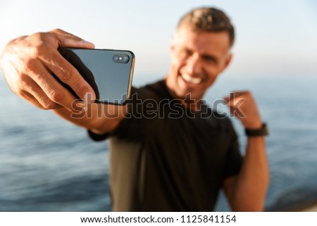Confident handsome shirtless sportsman taking a selfie while standing at the beach and flexing biceps