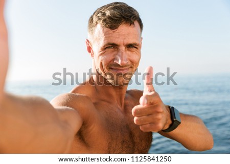 Smiling handsome shirtless sportsman taking a selfie and pointing finger at camera while standing at the beach