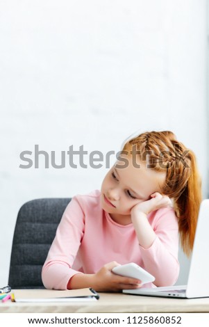 bored redhead kid looking away while using smartphone and laptop