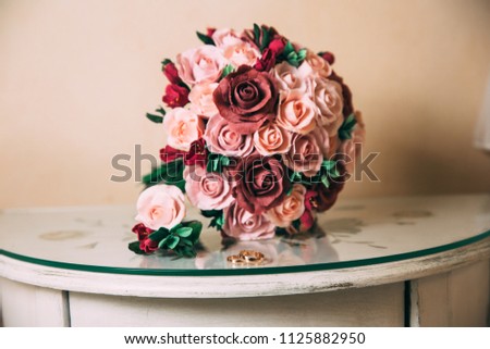 The gold rings of the newlyweds and the groom's boutonniere lie on a wooden vintage table. A wedding bouquet of red and pink roses lies on the background. The concept of wedding floristry and jewelry