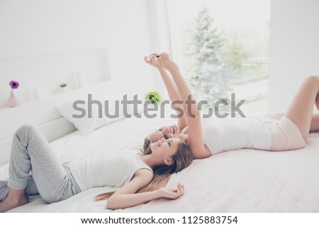 Pajama babysitter fashion style social network blogger concept. Two pretty cheerful excited joyful cute lovely charming girls using telephone making self picture lying on bed in white light room