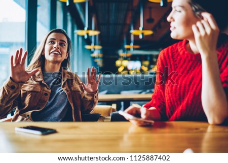 Two happy hipster girls discussing ideas for attracting followers for shared blog in social networks indoors, successful female users communicated at comfortable cafeteria while waiting friend Royalty-Free Stock Photo #1125887402