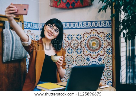 Positive female teenager enjoying tea time taking images on front mobile camera, cheerful woman sitting near bohemian wall and making video for blog using multimedia application on cellular phone