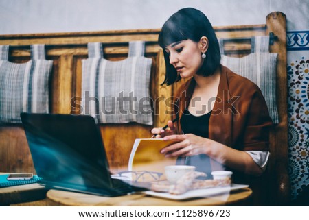 Clever intelligence female writer searching information for creating new best seller using modern laptop device and literature during free time at cafeteria, concept of education and knowledge