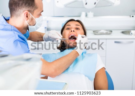 Diligent friendly dentist is treating woman patient which is sitting in dental chair in clinic.