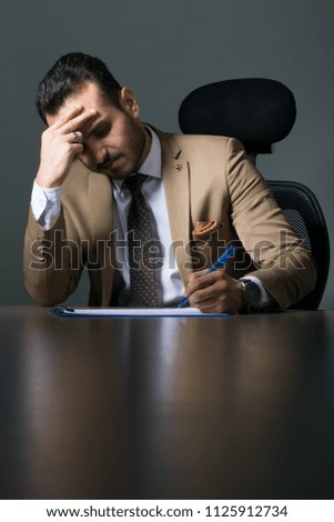 Concentrated young businessman writing documents at office desk, on a grey background.