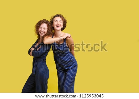Studio shot of two Caucasian sisters with same curly hair, wearing similar blue stripped overall, smiling happily, looking at camera, posing at yellow blank wall, standing to each other closely