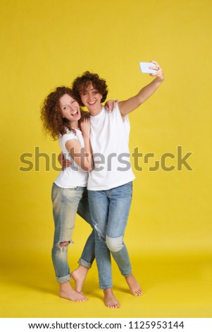Vertical shot of smiling female with cheerful expressions rejoice good mood, enjoy togetherness. Glad young best female friends or sisters make selfie by smart phone, broadly laught. Yellow background