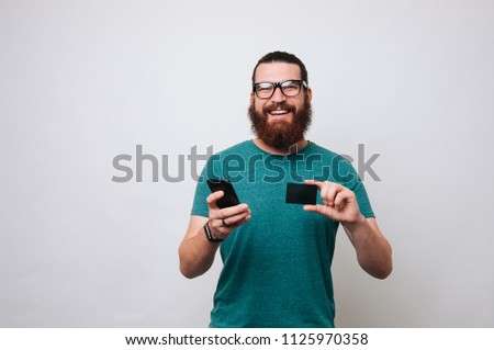 Photo of happy young bearded man isolated over white wall background holding mobile phone and credit card. Looking at the camera