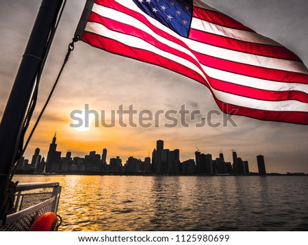A beautiful picture of an American Flag rippling in the wind over the Chicago skyline at sunset with building silhouettes and orange colored sun reflecting on the water of Lake Michigan.